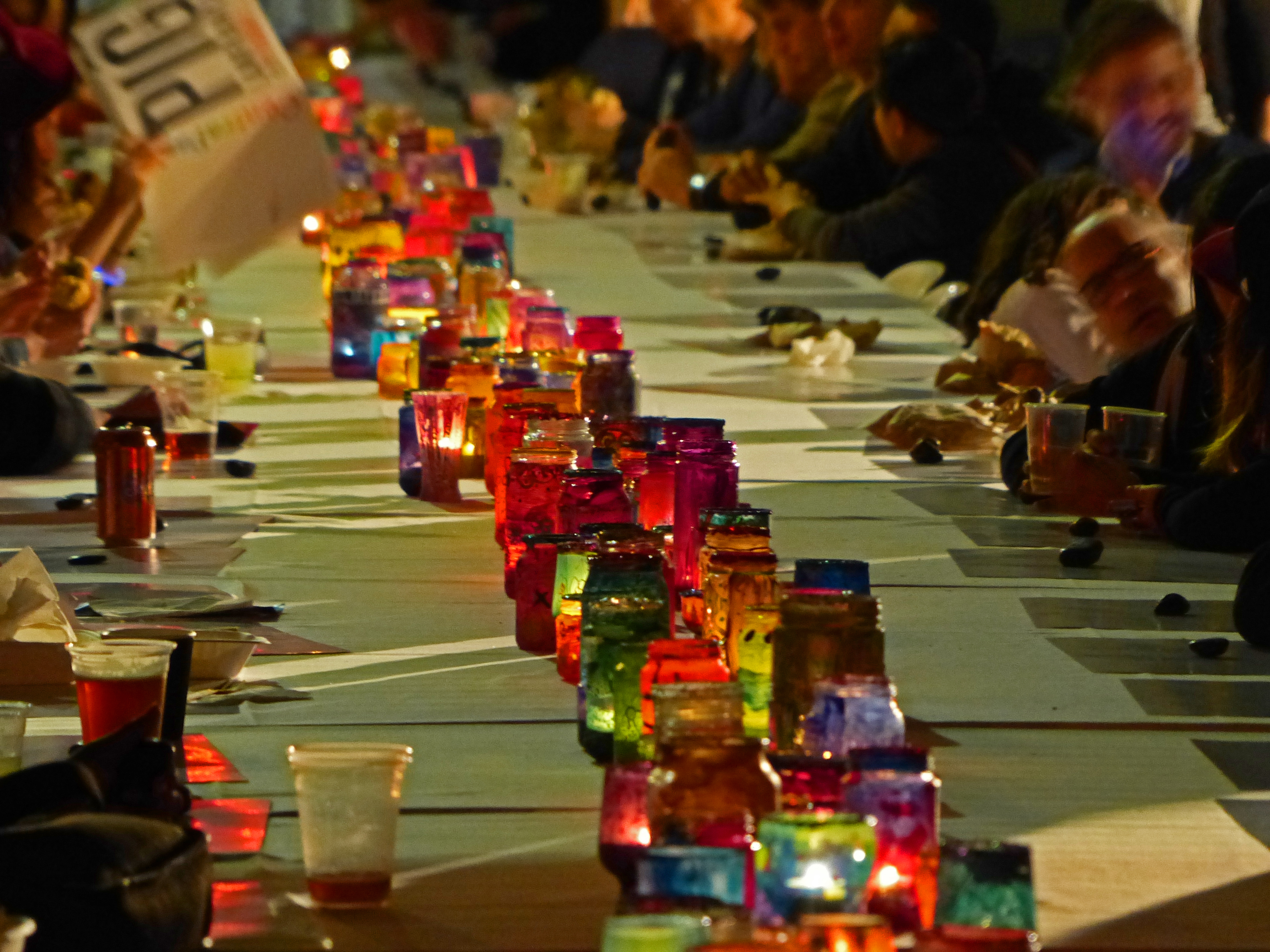 Bunte Gläser mit Teelichtern stehen auf der großen Essenstafel an der Menschen sitzen und essen